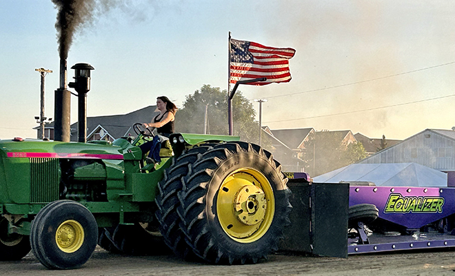 a woman driving a green tractor pulls a sled at the Out-of-Field Tractors, Semis and Pick-ups Pull