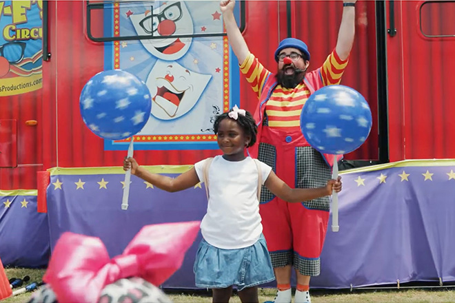 a girl spins balls on sticks while Lew-E the clown cheers her on