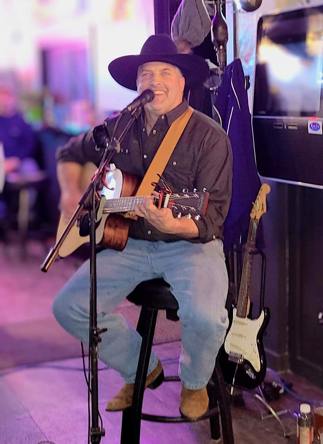 Grady Kruse sitting on a stool and strumming an acoustic guitar